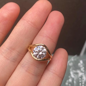 May include: A gold-colored ring with a large, round, clear gemstone set in a unique bezel design. The ring is held in a hand, with the text "GOLDENGLORY09 JEWELS" visible in the image.