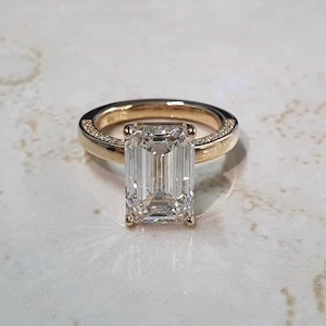 May include: A gold ring featuring a large, rectangular emerald-cut clear gemstone set in a four-prong mounting. The band is crafted from polished gold and includes a delicate row of small, sparkling pavé-set stones along the upper shank.
