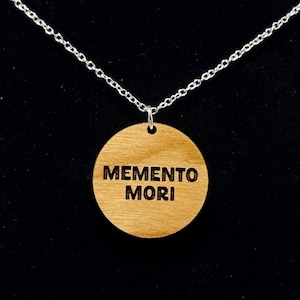 May include: A wooden pendant necklace with the words "MEMENTO MORI" engraved on a round, light brown wooden disc. The pendant hangs from a delicate silver chain against a black background.