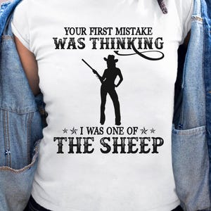 K&ouml;nnte beinhalten: Wei&szlig;es T-Shirt mit dem Text "YOUR FIRST MISTAKE WAS THINKING" &uuml;ber einer Silhouette einer Person, die ein Gewehr h&auml;lt. Darunter steht "I WAS ONE OF THE SHEEP".
