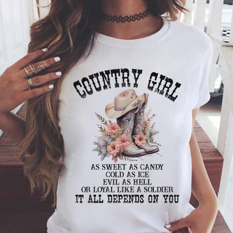 K&ouml;nnte beinhalten: Wei&szlig;es T-Shirt mit dem Text "COUNTRY GIRL" &uuml;ber einer Grafik von Cowboyhut und -stiefeln, umgeben von Blumen. Das Shirt zeigt auch den Text "AS SWEET AS CANDY COLD AS ICE EVIL AS HELL OR LOYAL LIKE A SOLDIER IT ALL DEPENDS ON YOU."
