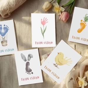 Puede incluir: Juego de cinco tarjetas de felicitación de Pascua con ilustraciones en acuarela. Los diseños incluyen una zanahoria de huella, una flor de huella de mano, un conejito de huella, un globo aerostático de huella y un pájaro de huella de mano. Cada tarjeta tiene el texto "Frohe Ostern".