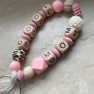 Könnte beinhalten: Ein Schlüsselanhänger mit Perlen, auf dem die Wörter "LOL" und "MOM" in Holzbuchstabenblöcken geschrieben sind. Der Schlüsselanhänger hat rosa, beige und leopardenmusternde Perlen, eine weiße Quaste und einen silbernen Metallring.