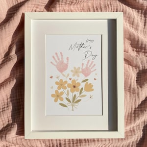 Könnte beinhalten: Ein gerahmter Druck mit dem Text "Happy Mother's Day". Das Kunstwerk zeigt zwei Handabdrücke über einem Strauß gelber und pfirsichfarbener Blumen. Der Rahmen ist weiß und der Hintergrund ist ein rosa Stoff.