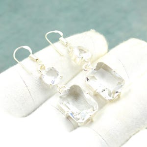 Puede incluir: Un par de pendientes de plata con piedras preciosas claras y facetadas. Cada pendiente presenta un gancho, una piedra redonda y una piedra rectangular más grande. Las piedras están engastadas en metal plateado.