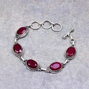 Puede incluir: Una pulsera de plata con cinco piedras preciosas de rubí rojo oscuro de forma ovalada, engastadas en marcos de metal trenzado. La pulsera incluye un cierre de barra y tres anillos de ajuste circulares en un extremo que unen los engastes.