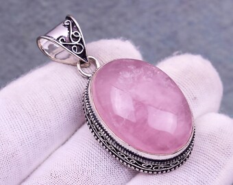 May include: A silver pendant with an oval, light pink gemstone. The gemstone is set in a decorative silver frame with an ornate bail. The pendant is displayed on a white surface.
