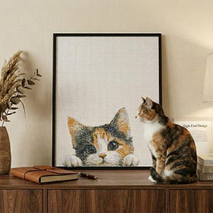 Puede incluir: Un cuadro negro con una obra de arte de un gato calicó. Un gato calicó real está sentado al lado. Un jarrón con plantas secas, libros y una lámpara también están sobre una superficie de madera.