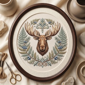 May include: A framed cross-stitch featuring a moose head with large antlers, surrounded by a wreath of green and blue foliage. The artwork is set against a cream-colored background and framed in a dark brown wooden frame. The image also includes embroidery floss and scissors.