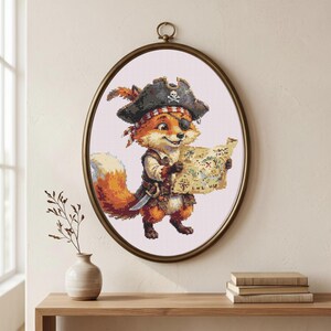 May include: A framed cross-stitch artwork depicts a cartoon fox dressed as a pirate, wearing a hat with a skull and crossbones, an eye patch, and holding a treasure map. The fox is in a gold oval frame, with a small vase and books on a wooden surface below.