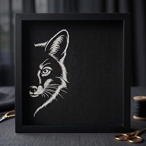 May include: A square black frame containing a piece of black fabric with a white embroidered design of the left side of a fox's face. The embroidery features detailed stitching of the fox's eye, ear, snout, and whiskers against the dark background.