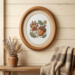May include: An oval wooden frame containing a cross-stitch embroidery of a brown animal paw print and a brown acorn, surrounded by small green and yellow oak leaves on a white fabric background, displayed as a piece of wall art.