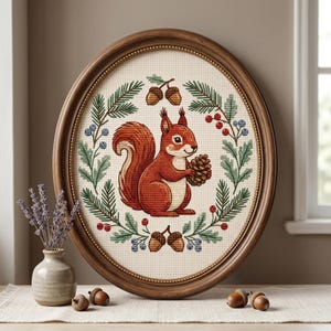 May include: Oval framed cross-stitch art depicting a red squirrel with a pinecone, encircled by foliage, berries, and acorns. The artwork is set against a cream backdrop and framed in a dark wood oval frame. A small vase of lavender is placed nearby.