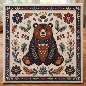 May include: A square cross-stitch embroidery piece featuring a central brown bear surrounded by a symmetrical pattern of flowers, oak leaves, pine branches, and red berries, all enclosed within a dark blue decorative border on cream fabric.