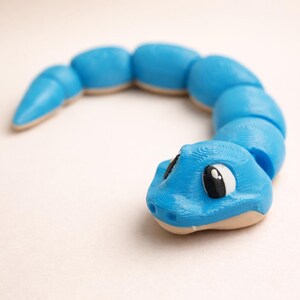 May include: A bright blue articulated snake toy with a segmented body and a smooth, curved shape. The head features large, white, and black eyes with a visible 3D-printed texture throughout the surface, ending in a tapered tail section.