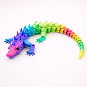 May include: A multi-colored, articulated lizard figurine featuring a gradient color scheme that transitions from pink and purple at the head to blue, green, yellow, and orange along its segmented body and long, curved tail with sharp dorsal spikes.