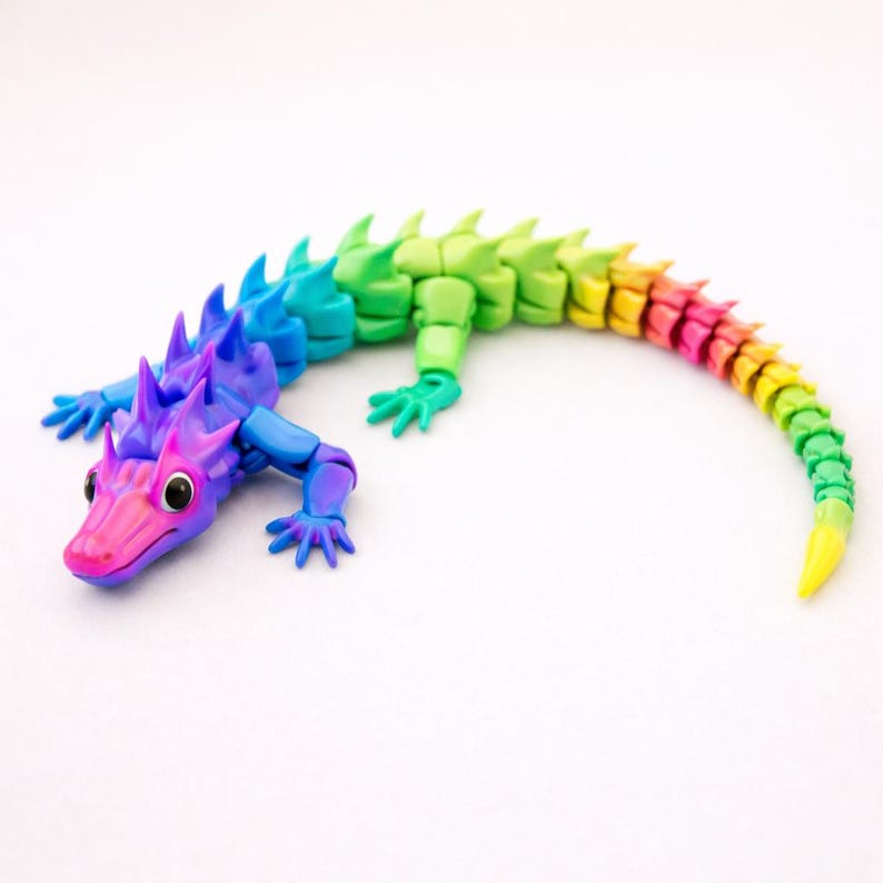 May include: A multi-colored, articulated lizard figurine featuring a gradient color scheme that transitions from pink and purple at the head to blue, green, yellow, and orange along its segmented body and long, curved tail with sharp dorsal spikes.