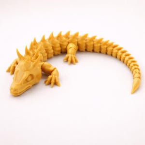 May include: A mustard yellow articulated lizard figurine featuring a segmented body with sharp, pointed spikes running along the back and tail. The creature has a detailed head with defined eye sockets and four limbs with clawed feet, all in a solid color.