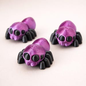 May include: Three small spider figurines with metallic purple bodies and black legs. Each spider features a rounded abdomen and a head with large, prominent black eyes. The figures have a textured surface finish consistent with 3D printing.