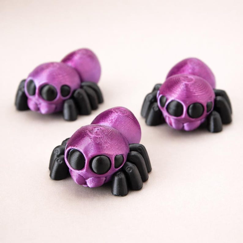 May include: Three small spider figurines with metallic purple bodies and black legs. Each spider features a rounded abdomen and a head with large, prominent black eyes. The figures have a textured surface finish consistent with 3D printing.