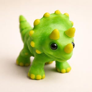 May include: A small, bright green triceratops dinosaur figurine with a textured surface. The creature features three prominent yellow horns on its head, a large frill with yellow rounded spikes along the edge, and yellow accents on its feet.