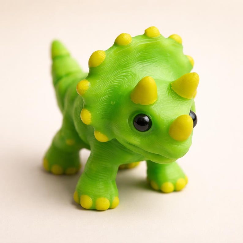 May include: A small, bright green triceratops dinosaur figurine with a textured surface. The creature features three prominent yellow horns on its head, a large frill with yellow rounded spikes along the edge, and yellow accents on its feet.