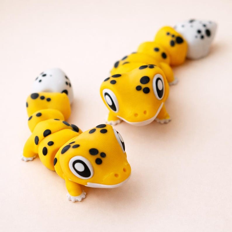 May include: Two articulated lizard figurines featuring a bright yellow body with black spots and large white eyes with black pupils. Each lizard has a segmented body structure and a white tail tip, showcasing a smooth matte finish and rounded features.