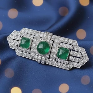 Peut inclure: Broche argentée ornée de trois émeraudes et de nombreux petits diamants. La broche rectangulaire présente une émeraude centrale circulaire et deux émeraudes carrées. Les pierres sont serties dans un design géométrique détaillé.