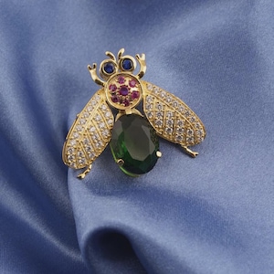 May include: A gold-toned brooch in the shape of a fly, featuring a large green oval gemstone for the body. The wings are adorned with clear crystals, and the head has blue and red gemstones. The brooch is set against a blue fabric.