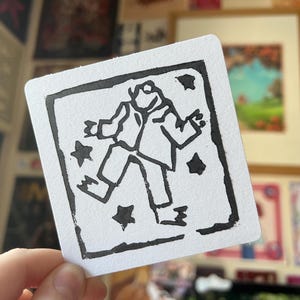 May include: A white square card with a black linocut print of a figure with outstretched arms and legs, and black star shapes. The figure is wearing a suit. The card is held in a hand.
