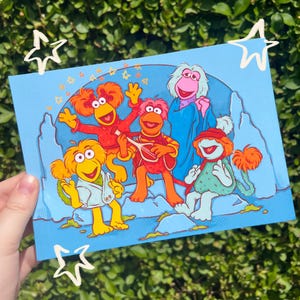 May include: A rectangular postcard featuring a colourful illustration of five Fraggles against a blue background. The characters include orange, pink, and light blue creatures with distinct hair, one playing a stringed instrument, surrounded by small yellow stars.