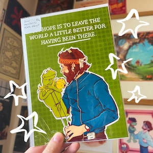 May include: A rectangular art print in a clear plastic sleeve featuring an illustration of a bearded person wearing headphones and a headband, holding a green frog puppet. The text reads, My hope is to leave the world a little better for having been there.