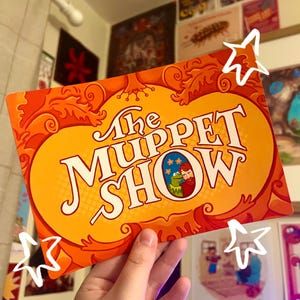 May include: A rectangular orange and yellow card featuring the text The Muppet Show in a stylised font. The design includes swirling leaf-like patterns and a small circular illustration of Kermit the Frog and a man with a beard against a blue star background.