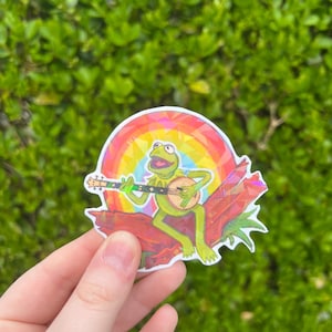 May include: A sticker depicting a green frog playing a banjo beneath a rainbow. The frog is perched on a red branch with green leaves. The background is a vivid green.