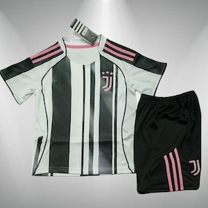 Pode incluir: Uma camisa de futebol listrada em branco e preto e calções pretos combinando. A camisa tem detalhes em rosa nos ombros e um logotipo da equipe no peito. Os calções têm listras rosa e um logotipo da equipe. Uma etiqueta está anexada à camisa.