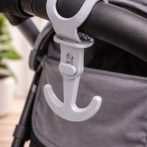 May include: A gray plastic stroller hook attached to a black stroller handle. The hook has a curved top and a lower hook shape, designed for hanging items. The stroller's gray fabric and black frame are visible in the background.