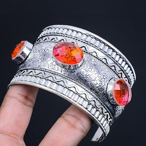 May include: Silver cuff bracelet featuring three orange-red gemstones. The bracelet has a detailed floral and geometric design. The gemstones are oval-shaped and set in silver bezels. The bracelet is displayed against a dark backdrop.