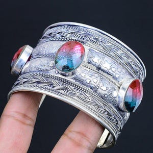 Könnte beinhalten: Ein breites silbernes Armreifarmband mit drei oval geschliffenen Edelsteinen. Die Edelsteine zeigen einen Farbverlauf aus Rot, Blau und Grün. Das Armband hat aufwendige geflochtene und geätzte Details.