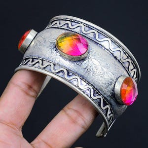 May include: Silver cuff bracelet featuring three oval, multi-coloured gemstones. The bracelet has a detailed, etched design and a wavy border. The gemstones are shades of red, orange, and yellow.