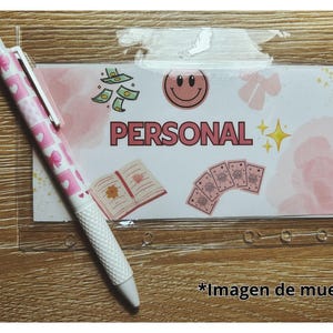 Puede incluir: Un sobre de plástico transparente con la palabra "PERSONAL" en rosa, decorado con dinero, una carita sonriente y otros gráficos. Un bolígrafo rosa con un patrón de corazones está sobre la superficie de madera.