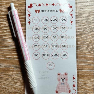 Può includere: Una penna rosa e bianca è appoggiata accanto a una scheda di sfida di risparmio. La scheda presenta un'illustrazione di orsetto e cerchi con importi da 5 a 20 euro. Il testo sulla scheda recita "RETO 200 €" e "RUMBO AL AHORRO".