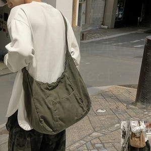 May include: Olive green canvas shoulder bag with a large front pocket and a long strap. The bag is worn over the shoulder. The bag is a casual style.