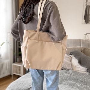 May include: A beige tote bag with cream-coloured straps and two exterior pockets. The bag has a rectangular shape and is held by a person wearing a grey sweatshirt and blue jeans. The bag has two buttons on the front.