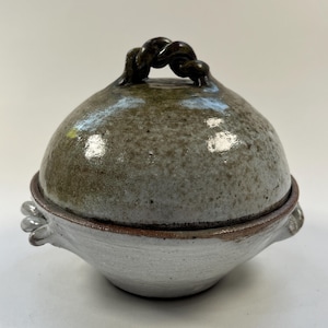 May include: A handmade ceramic lidded bowl. The bowl has a white base with a speckled, gray-green lid. The lid features a dark brown, twisted handle. The bowl has small, decorative handles on either side. A unique piece for kitchen or home decor.