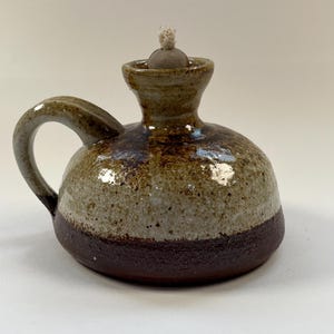 May include: A handmade ceramic oil lamp with a handle. The lamp has a rounded body with a dark brown base, a speckled tan middle, and a brown top. A wick sits atop the lamp. The lamp is designed for use with oil.
