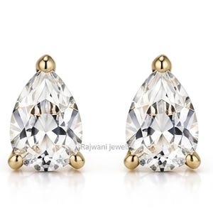 Peut inclure: Une paire de pierres précieuses transparentes taille poire montées sur des clous en métal doré. Chaque pierre est fixée par trois petites griffes dorées aux extrémités. Le texte Rajwani jewels est visible en gris sur fond blanc.