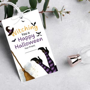 Op de afbeelding: Een witte Halloween-cadeaulabel met de tekst "Witching You a Happy Halloween". De tag bevat illustraties van een heksenhoed, bezem en benen met paarse en zwart gestreepte kousen. Inclusief een wit touwtje om op te hangen.