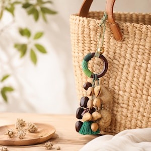 May include: A woven straw tote bag with a brown leather handle. A decorative wooden ring with green and brown fabric accents hangs from the handle, adorned with brown and tan beads and a green tassel.