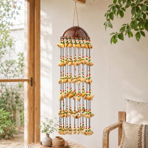 May include: A hanging wind chime with a coconut shell top and multiple strands of natural-colored shells. The strands are adorned with red, green, and yellow beads. The wind chime hangs from a brown string.
