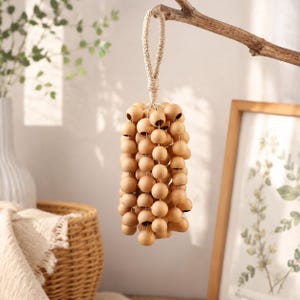 May include: A decorative hanging ornament made of light brown wooden beads. The beads are strung together in a cascading pattern and suspended from a natural fiber loop. The ornament is hanging from a tree branch.